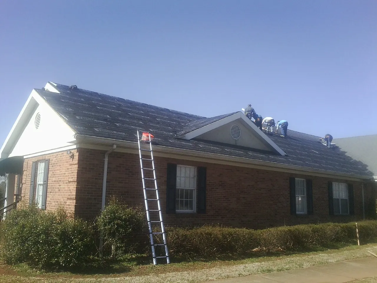 Crew tearing shingles off a brick ranch roof with a ladder set — Metal Roof Repair work by our crew in Byfield