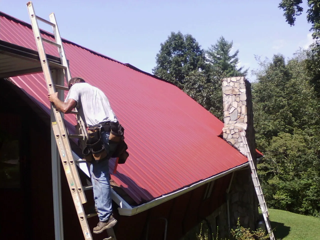 Roofer on a ladder beside a red metal roof and stone chimney — Soffit & Fascia Repair by Roofing Brothers in Byfield