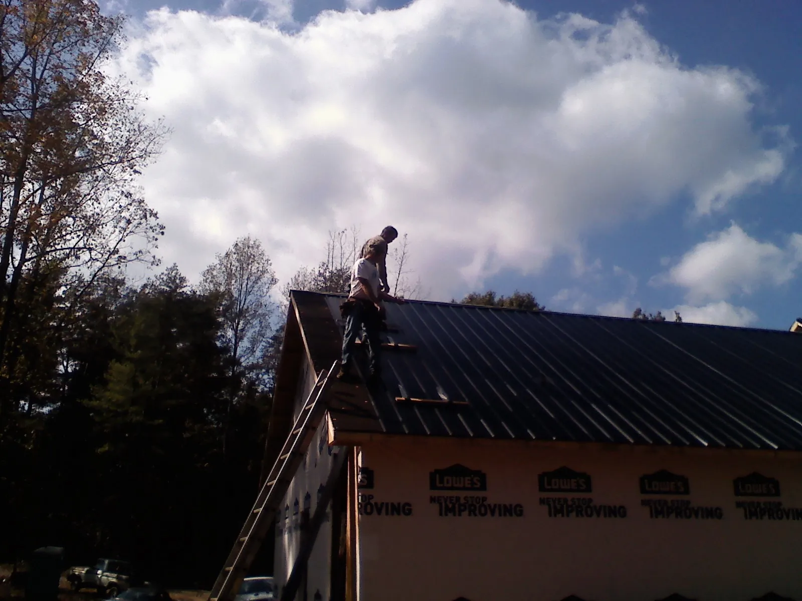 Roofer fastening dark metal panels high against a cloudy sky, from a Roof Inspection job in Byfield